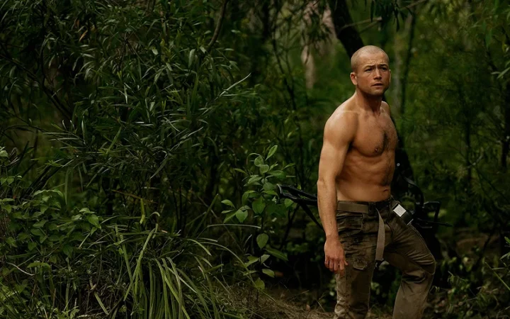 Taron Egerton sem camisa e armado com uma besta na floresta em cena de O Jogo do Predador, crítica e suspense.