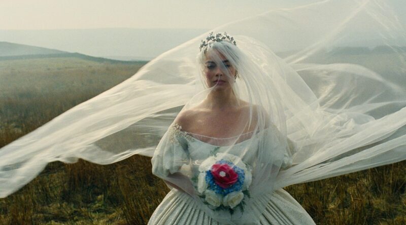 Margot Robbie como Catherine Earnshaw vestida de noiva com véu longo ao vento em cenário de campo de O Morro dos Ventos Uivantes.