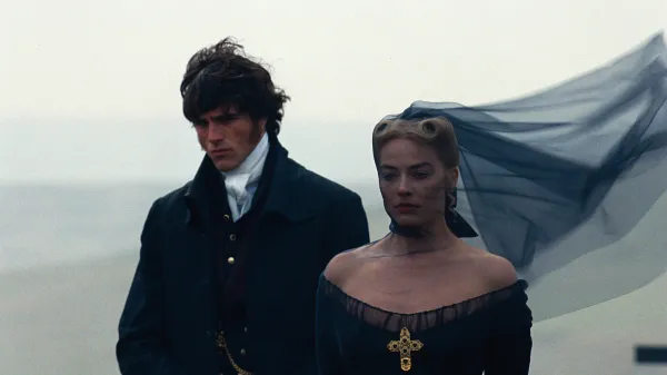 Margot Robbie como Catherine usando vestido preto com cruz dourada e Jacob Elordi como Heathcliff em cenário aberto de O Morro dos Ventos Uivantes