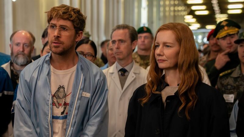 Ryan Gosling como Ryland Grace e Sandra Hüller como Eva Stratt caminhando por um corredor ladeado por militares e cientistas em Devoradores de Estrelas.