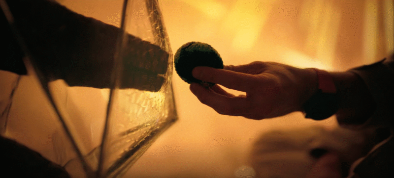 Detalhe da mão de Ryland Grace tocando a redoma de vidro do alienígena Rocky, em um momento de conexão emocional sob luz âmbar em Devoradores de Estrelas.