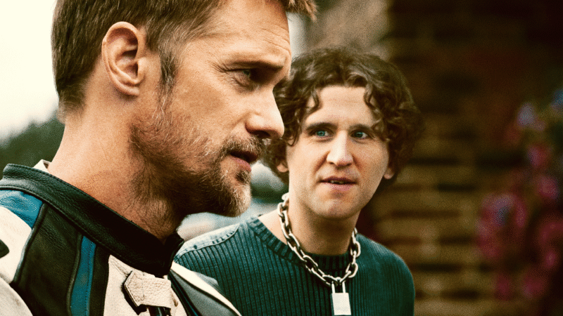 Alexander Skarsgård de perfil em primeiro plano com expressão séria usando jaqueta de motociclista, enquanto Harry Melling o observa ao fundo usando uma grande corrente com cadeado, ilustrando as novidades nos streamings.