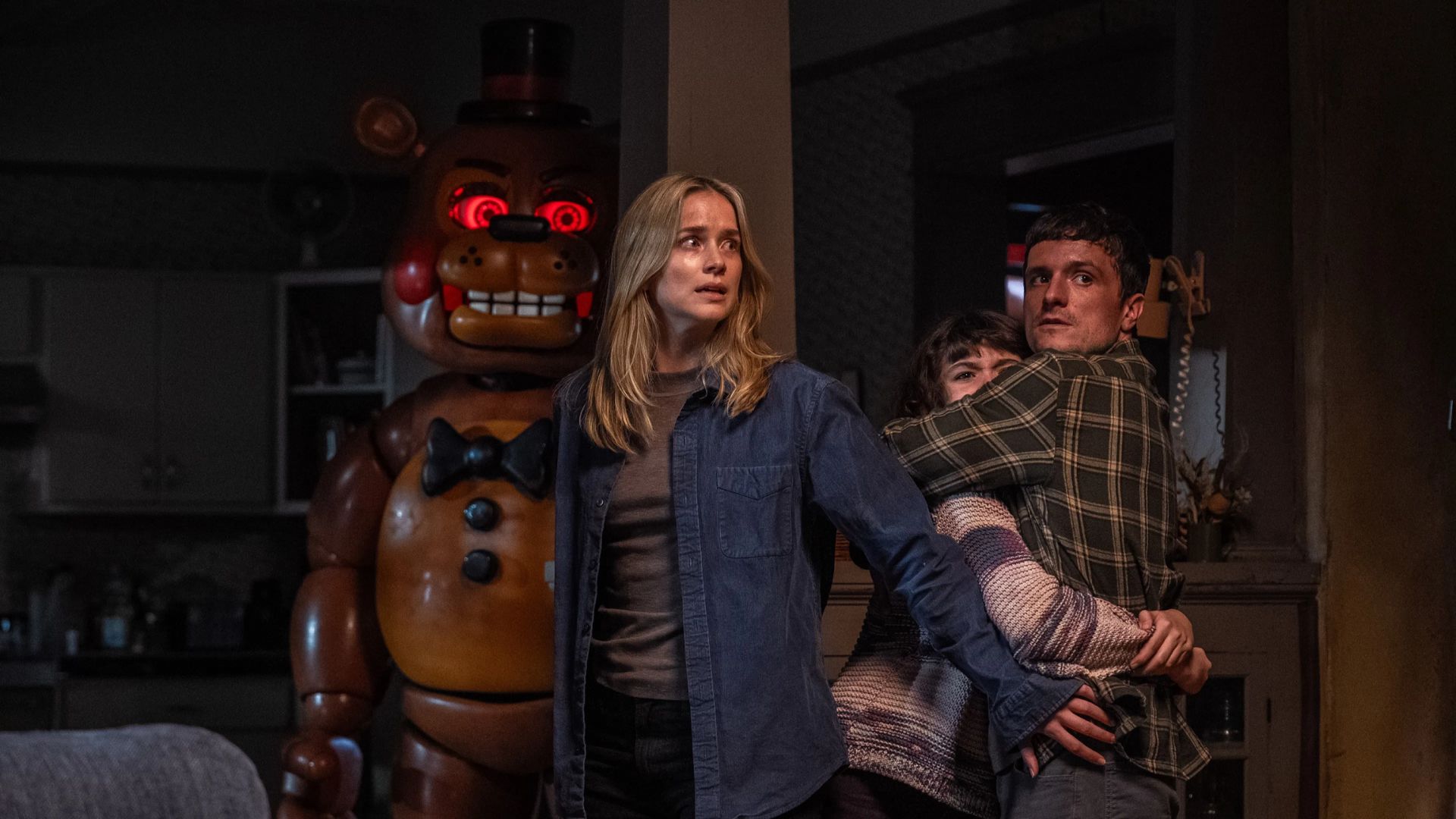 Elizabeth Lail com expressão assustada estende o braço protegendo Josh Hutcherson e Piper Rubio abraçados em um cômodo escuro, enquanto o urso animatrônico Toy Freddy surge ao fundo com olhos vermelhos brilhantes, ilustrando as novidades nos streamings.