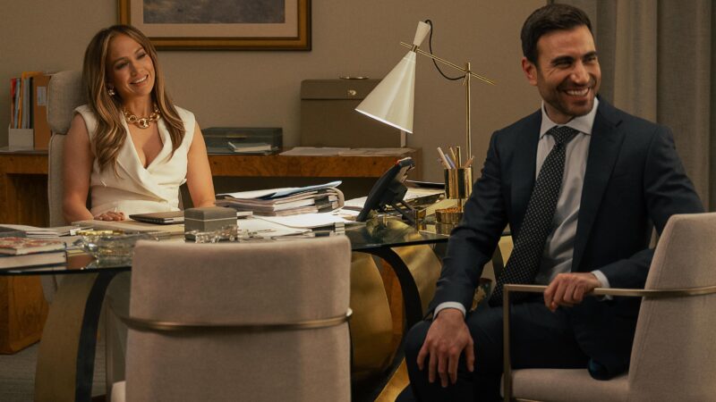 Jennifer Lopez sorri em sua mesa de escritório vestindo um colete branco estruturado enquanto Brett Goldstein, de terno e gravata, sorri olhando para o lado em primeiro plano, ilustrando as novidades nos streamings.