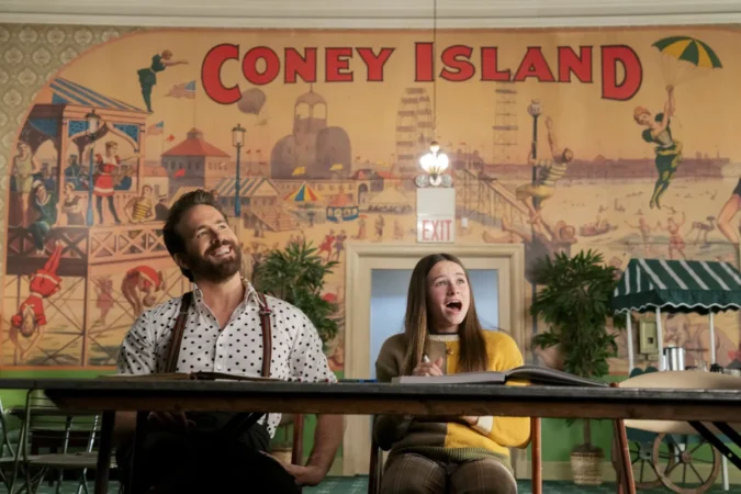 Ryan Reynolds e Cailey Fleming sentados em uma mesa sorrindo diante de um grande painel ilustrado de Coney Island no filme Amigos Imaginários, destaque das estreias nos streamings da semana.