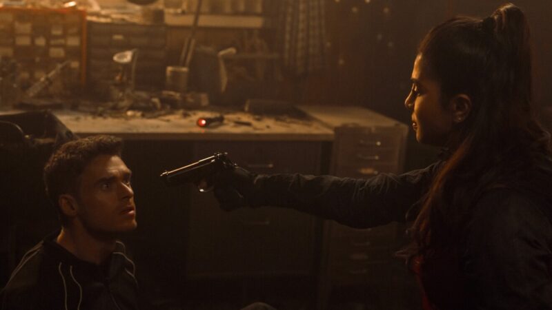 Cena de tensão na série Citadel onde Nadia Sinh aponta uma arma para Mason Kane em uma oficina escura.
