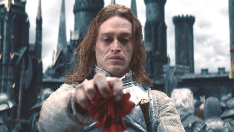 Caleb Landry Jones como Drácula de Luc Besson vestindo armadura e segurando flor vermelha.
