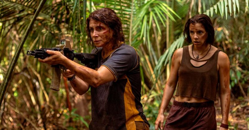 Giovanna Antonelli aponta um rifle tático para frente com olhar tenso enquanto Alice Wegmann observa ao fundo em uma floresta densa com terra e vegetação, ilustrando as novidades nos streamings.