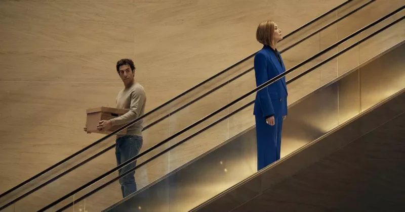 Sacha Baron Cohen segurando uma caixa de papelão e Rosamund Pike vestindo terno azul em escadas rolantes na comédia Primeiro as Damas, destaque das estreias nos streamings da semana.
