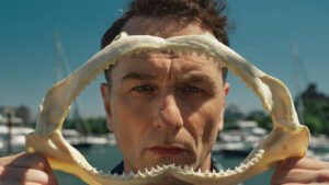 Ator Matthew Rhys segurando uma mandíbula de tubarão em cena de O Segredo de Widow's Bay.