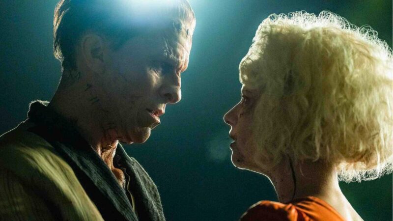 Christian Bale com cicatrizes marcantes no pescoço e Jessie Buckley com cabelos loiros cacheados se encaram bem de perto em um close dramático de perfil, ilustrando o filme a noiva.