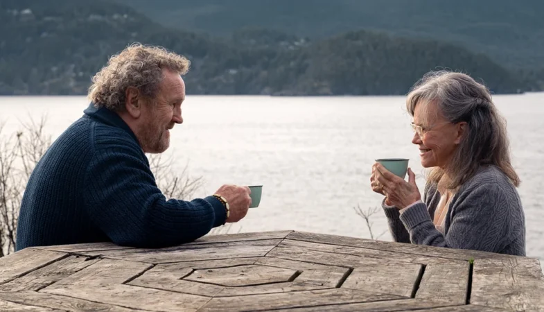 Colm Meaney e Sally Field conversando e tomando café em uma mesa de madeira ao ar livre, ilustrando o significado de Criaturas Extraordinariamente Brilhantes.