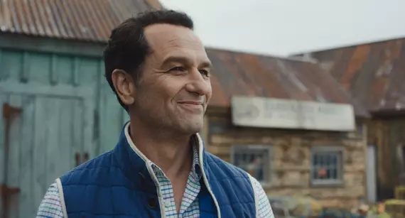 Ator Matthew Rhys sorrindo em cena externa na vila de O Segredo de Widow's Bay.