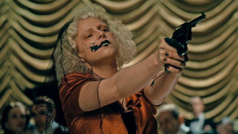 Jessie Buckley com batom preto borrado e véu escuro aponta um revólver para a frente com as duas mãos estendidas, ilustrando o filme a noiva.