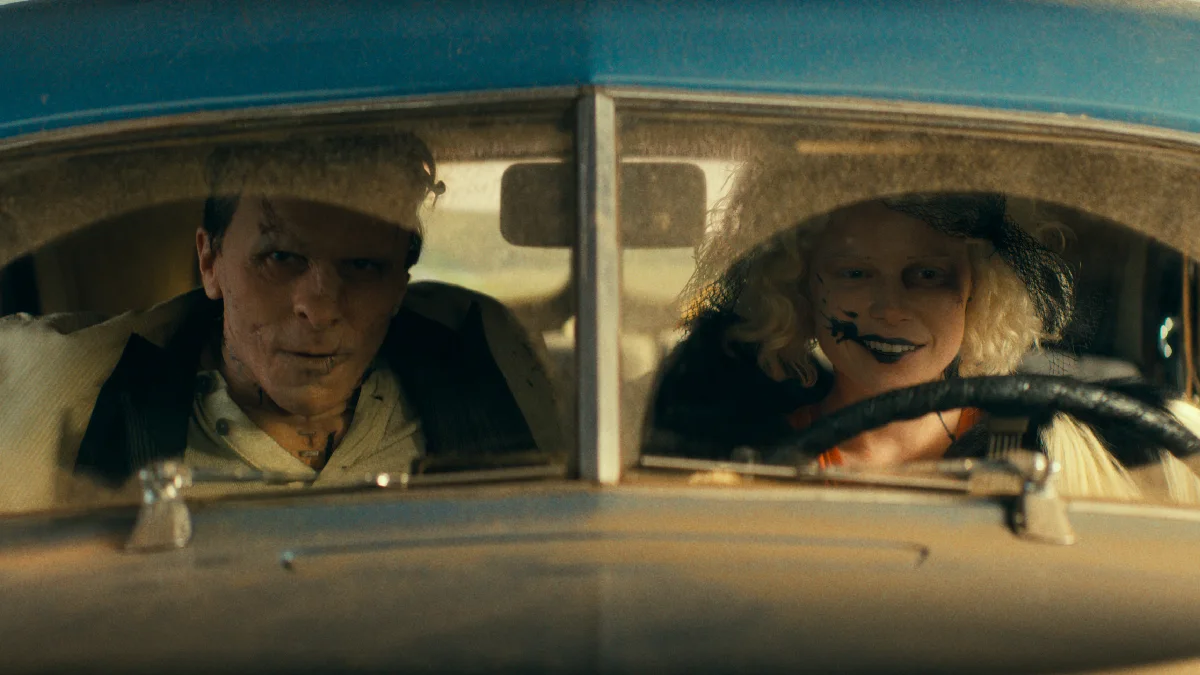 Christian Bale com cicatrizes de grampos na testa observa Jessie Buckley sorrindo com batom escuro borrado enquanto ela dirige um carro antigo azul, ilustrando o filme a noiva.