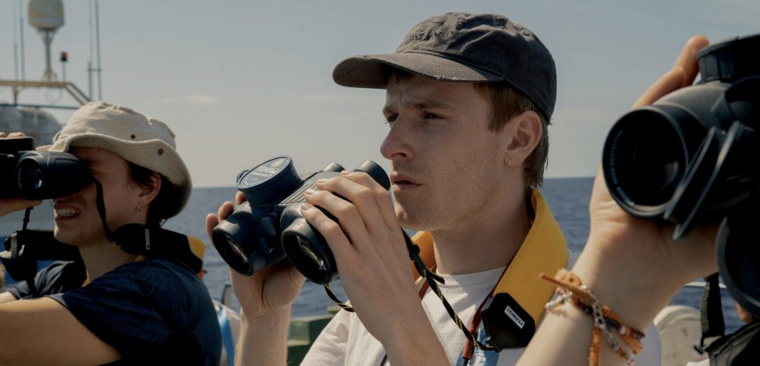 Louis Hofmann com binóculo em alto-mar na emocionante trama de 23.000 Vidas, destaque dos lançamentos de julho.