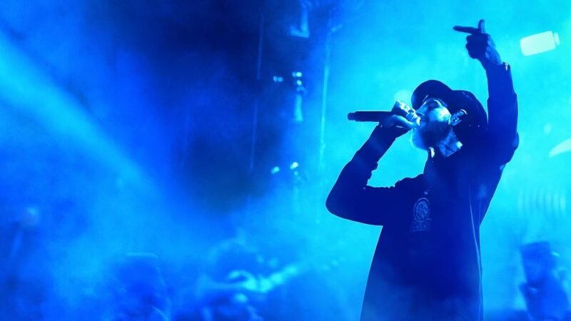 Silhueta de um cantor de boné e capuz no palco iluminado por fortes luzes azuis e fumaça, cantando em um microfone e apontando para o alto.