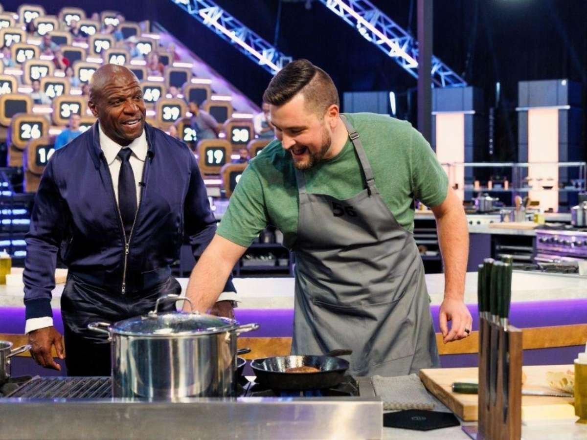 O apresentador Terry Crews, vestindo jaqueta escura e gravata, sorri e observa um cozinheiro de avental cinza mexendo em uma panela no fogão industrial de um estúdio de televisão.