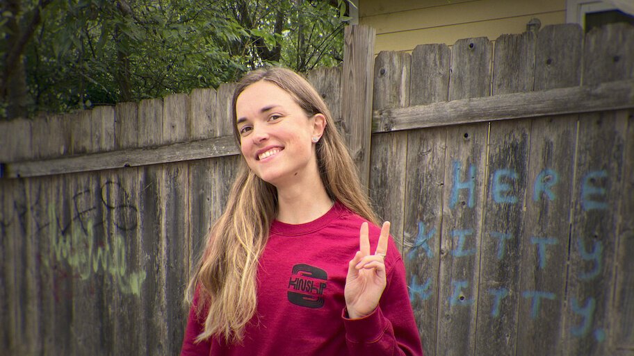Retrato de Moriah Wilson sorrindo de frente para a câmera e fazendo o sinal de paz com os dedos, vestindo uma blusa de moletom vinho diante de uma cerca de madeira com pichações, ilustrando o documentário da Netflix.