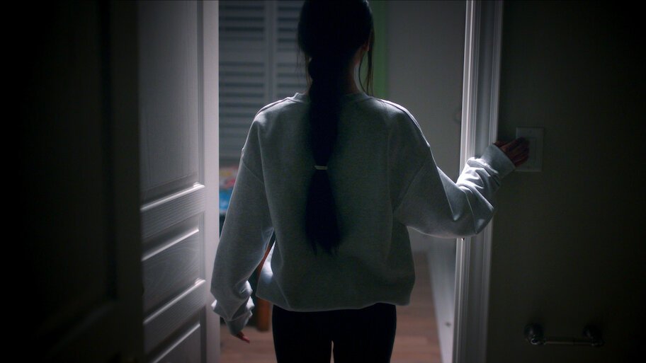 Mulher de costas e com cabelo preso em uma trança entra em um quarto escuro, estendendo a mão para o interruptor de luz. Uma das cenas do documentário de true crime da Netflix.