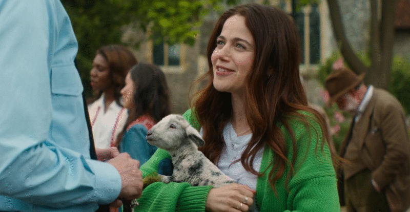 Molly Gordon em cena de "As Ovelhas Detetives" sorrindo enquanto segura um pequeno cordeiro malhado no colo e conversa com outra pessoa.