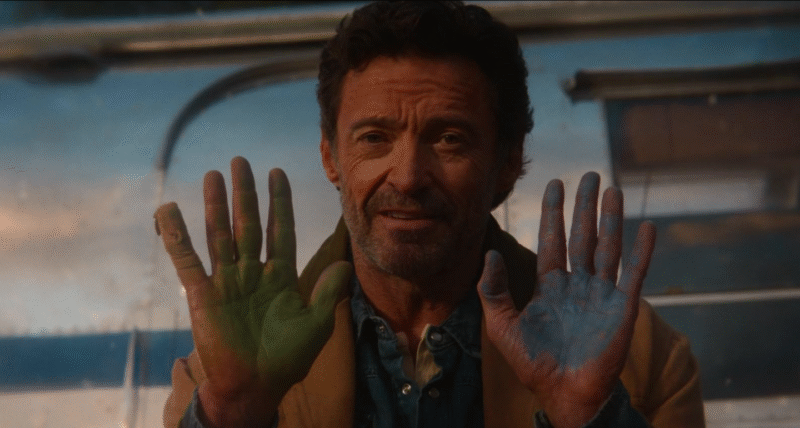 O ator Hugh Jackman sorrindo em cena de "As Ovelhas Detetives" enquanto mostra as palmas das mãos pintadas, uma de verde e a outra de azul.