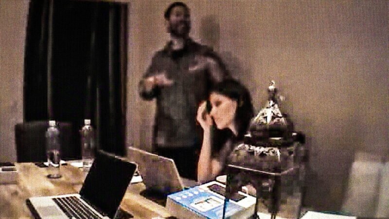 Registro de câmera de segurança mostrando Isabella Guzman sentada em frente a notebooks em uma mesa com Richard Rios de pé ao fundo, ilustrando a capa do post sobre documentários de true crime na Netflix.