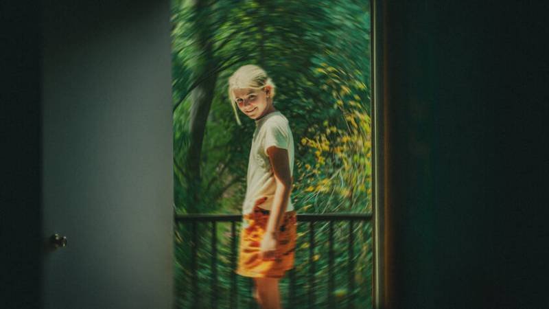 Foto desfocada e artística mostrando uma menina loira sorrindo de lado na sacada através de uma fresta de porta aberta, ilustrando o documentário de true crime da Netflix.