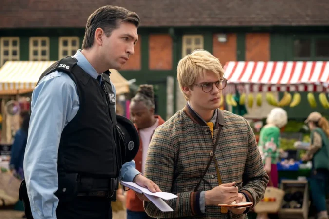 O ator Nicholas Braun como o policial Tim e Jonah Hauer-King como o jornalista em cena de "As Ovelhas Detetives", ambos segurando blocos de notas enquanto conversam em uma feira ao ar livre.