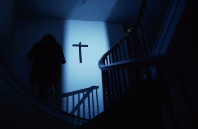 Silhueta de uma pessoa de costas em uma escada escura voltada para uma cruz preta na parede iluminada no filme Undertone.