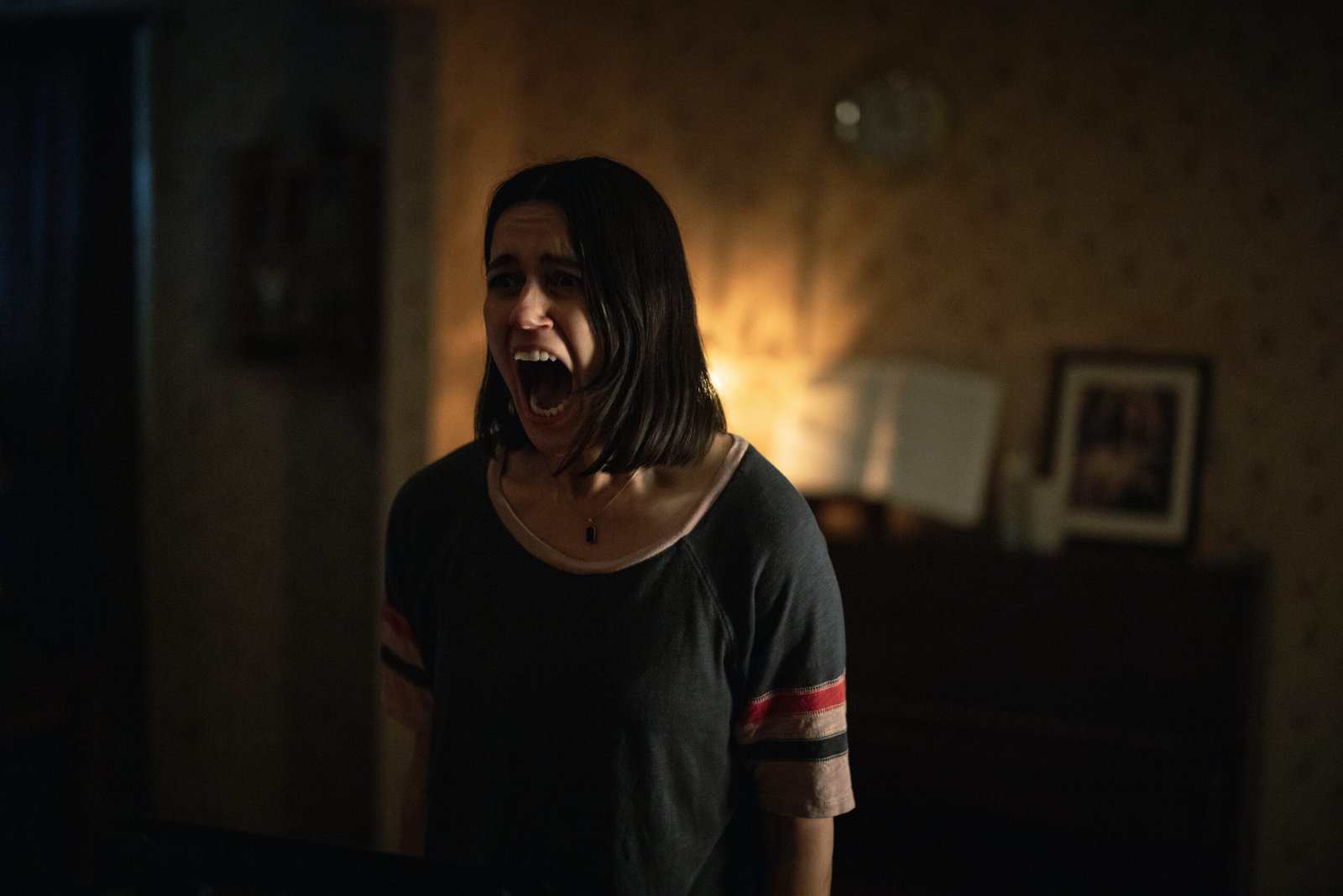 Mulher de cabelo curto gritando de pavor em uma sala escura e mal iluminada no filme Undertone.