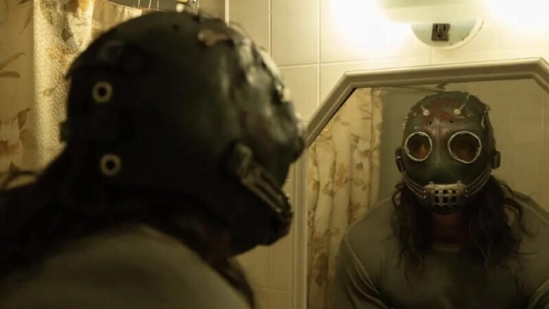 Homem com máscara assustadora de metal preta olhando o próprio reflexo no espelho do banheiro no filme Psycho Killer, destaque entre as estreias de agosto no streaming.