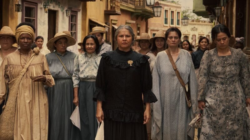 Grupo de mulheres de roupas de época caminhando sérias em uma rua de terra na série Cem Anos de Solidão, um dos destaques entre as estreias de agosto no streaming.