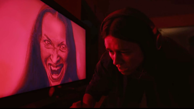Mulher com fones de ouvido olhando para o lado em uma sala com iluminação vermelha tensa ao lado de uma tela que exibe uma pintura digital assustadora no filme Undertone.
