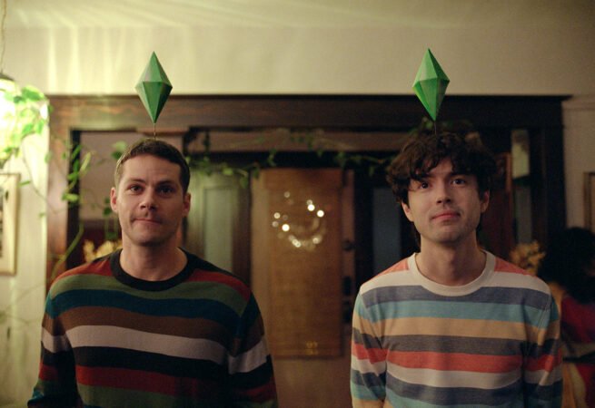 Dylan O'Brien e James Sweeney usando tiaras com o losango verde de The Sims no longa Twinless.