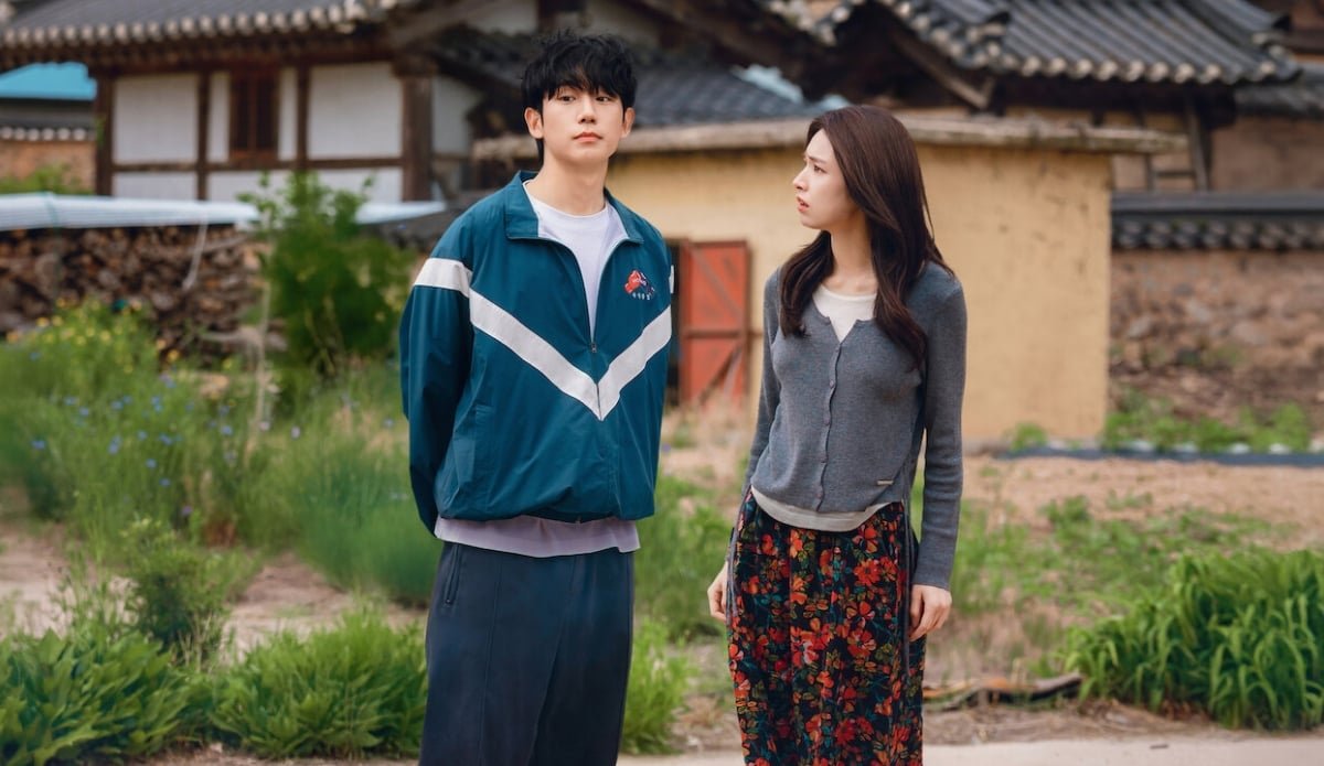 Um homem de jaqueta esportiva azul e uma mulher de saia floral conversando em uma vila rural no k-drama Um Grude de Amor, destaque entre as estreias de agosto no streaming.