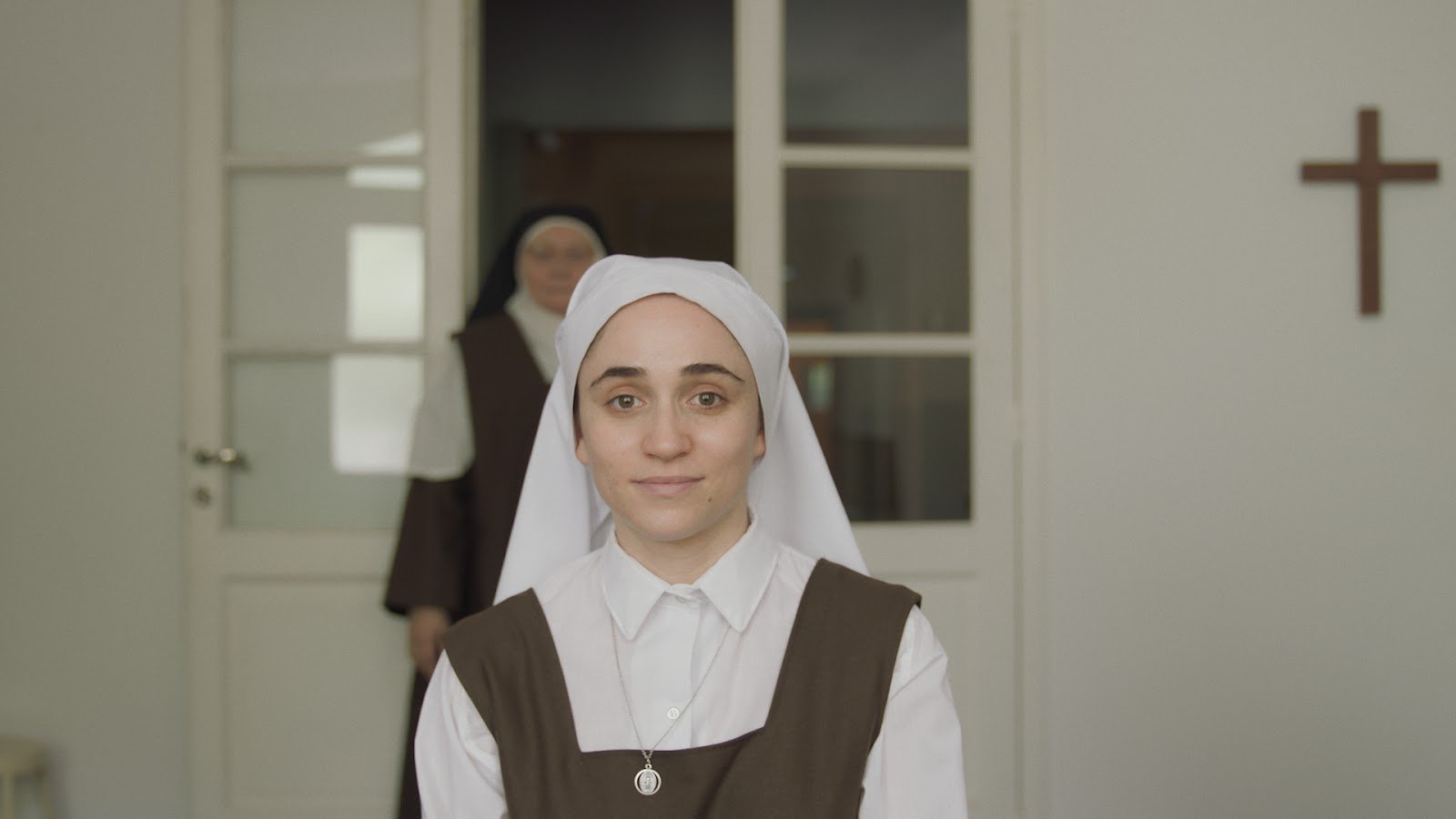 Clara com hábito religioso diante de uma porta, com outra freira ao fundo, em episódios de Confinada.