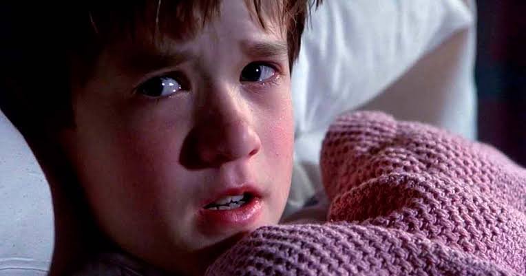 Haley Joel Osment como Cole Sear, assustado sob uma manta em O Sexto Sentido, uma das atuações de crianças em filmes e séries.