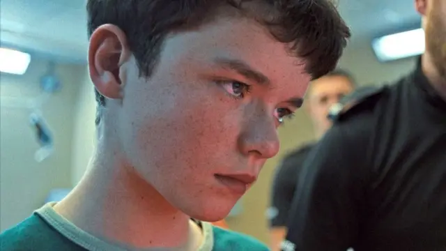 Owen Cooper como Jamie Miller, com expressão séria diante de policiais em Adolescência, destaque entre crianças em filmes e séries.