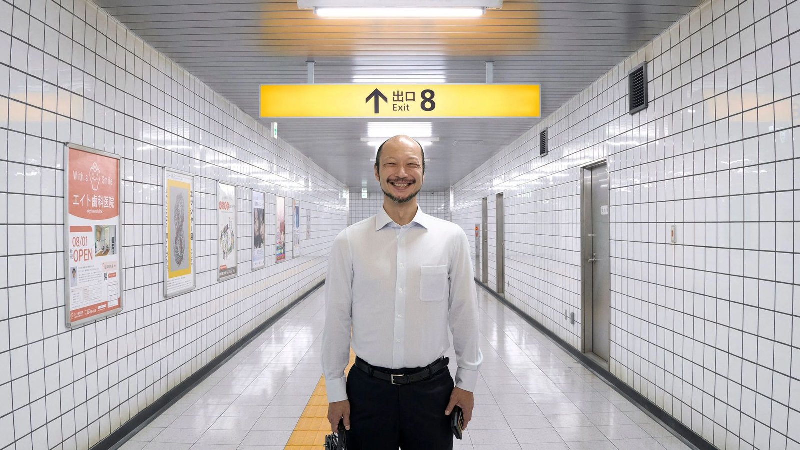 Homem sorridente sob a placa da saída 8 em um corredor subterrâneo no filme Exit 8, entre as novidades de outubro.