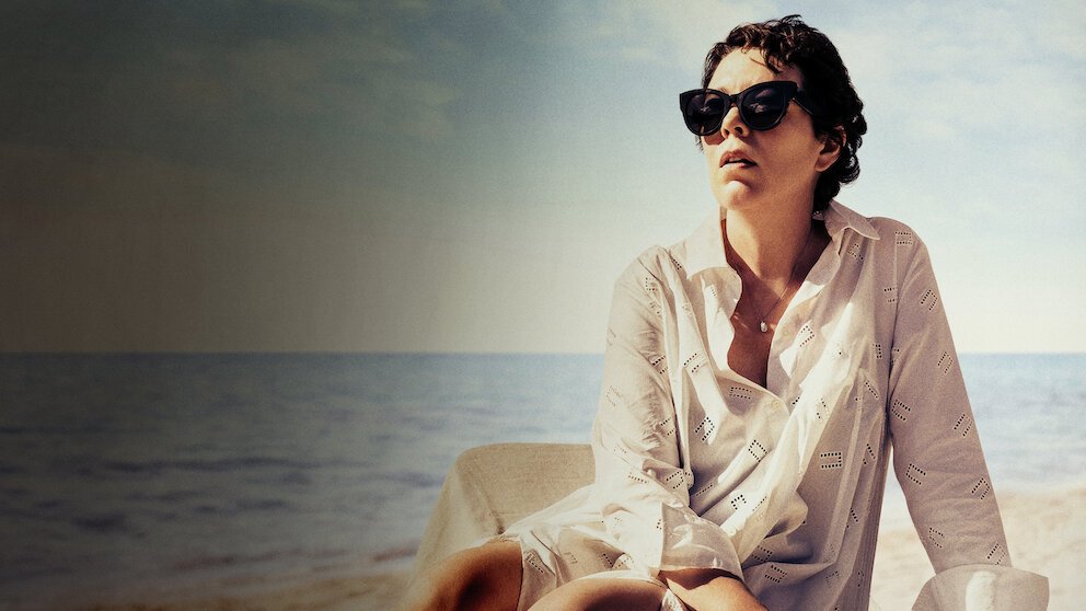 Olivia Colman como Leda sentada à beira-mar em A Filha Perdida, um dos filmes sobre o mar que exploram memória e maternidade.