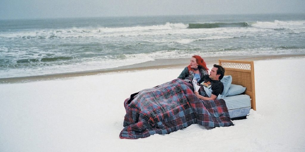 Jim Carrey e Kate Winslet deitados em uma cama na praia em Brilho Eterno de uma Mente sem Lembranças, da lista de filmes sobre o mar.