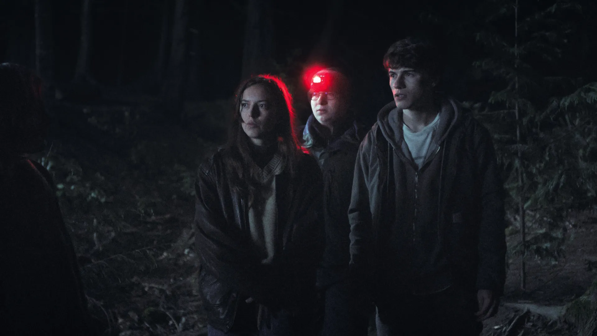 Sonny Cooke e dois jovens em uma floresta à noite, com uma lanterna vermelha, em cena da série Fingida (2026), do guia de episódios de Fingida.